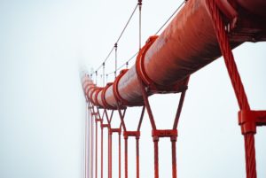 A red pipeline, upheld by a bridge of metal cables, fades into the white mist. Photo by JJ Ying on Unsplash