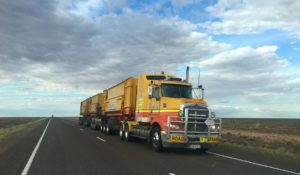 A lone truck or "road train" travels down an otherwise empty Australian road, beneath a blue and party cloudy sky