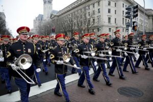 A line of trombonists leads a large military band in full regalia down a wide city street