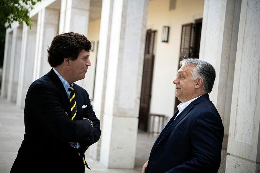 Tucker Carlson stands, arms crossed at his chest but pleased in front of a smiling Viktor Orban outside a collumned building in Hungary