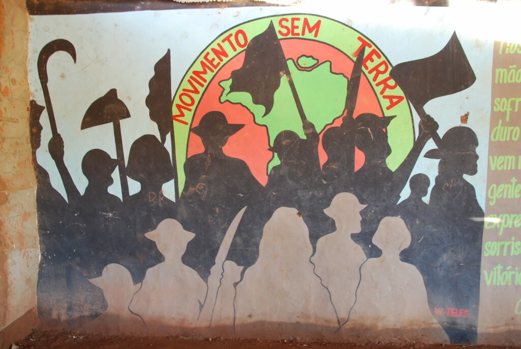 Silhouettes of demonstrators with flags and agricultural equipment in front of a representation of the globe bordered with the phrase Movimento Sem Terra painted on a wall