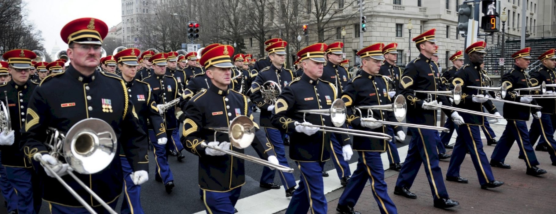 A line of trombonists leads a large military band in full regalia down a wide city street