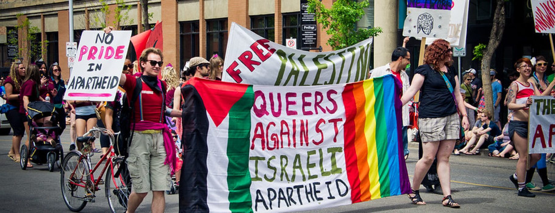 Two people carry a banner in a Pride march which says "Queers Against Israeli Apartheid" and which has on one side the flag of Palestine and on the othernd the rainbow flag. Behind them another banner reads "Free Palestine"