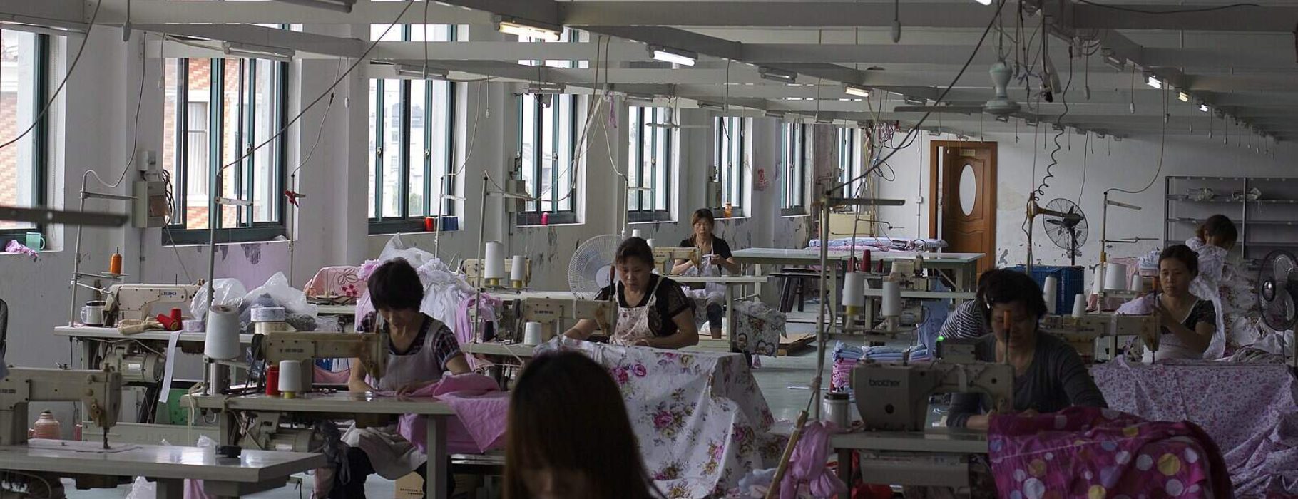 Poorly lit, partly empty silk factory with tired looking women hunched over their work.