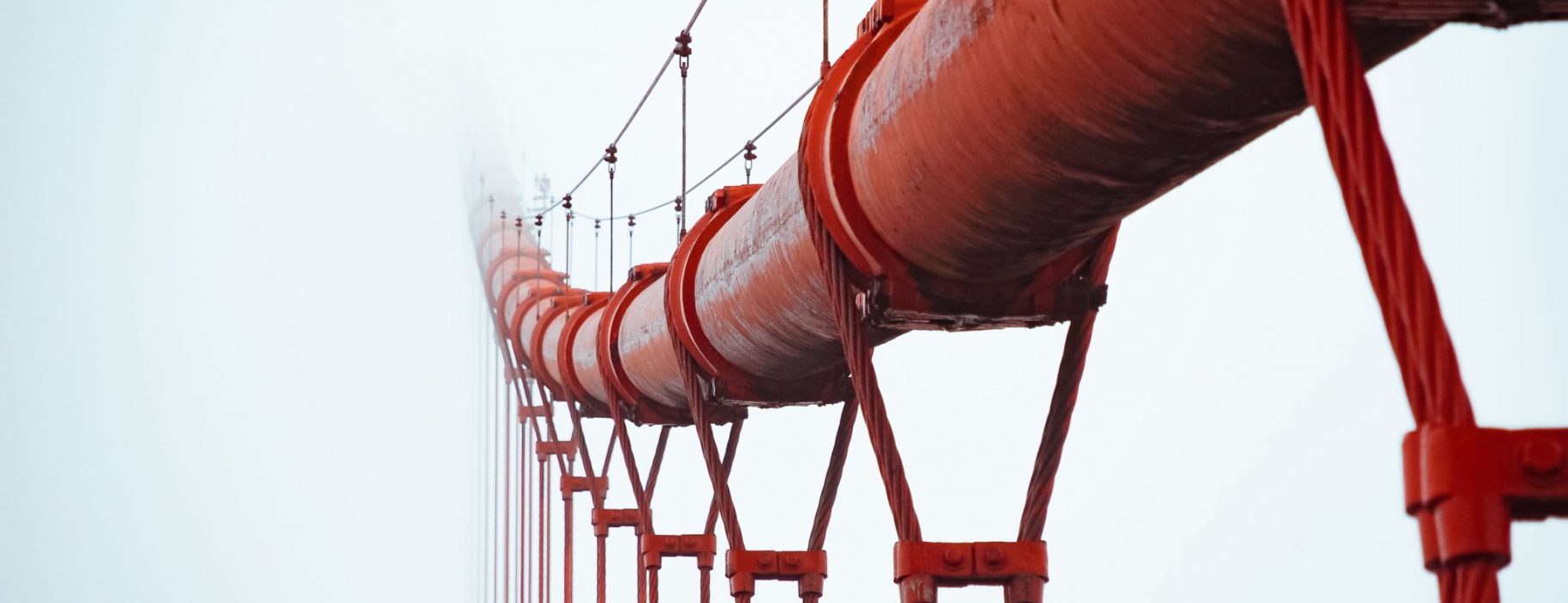 A red pipeline, upheld by a bridge of metal cables, fades into the white mist. Photo by JJ Ying on Unsplash