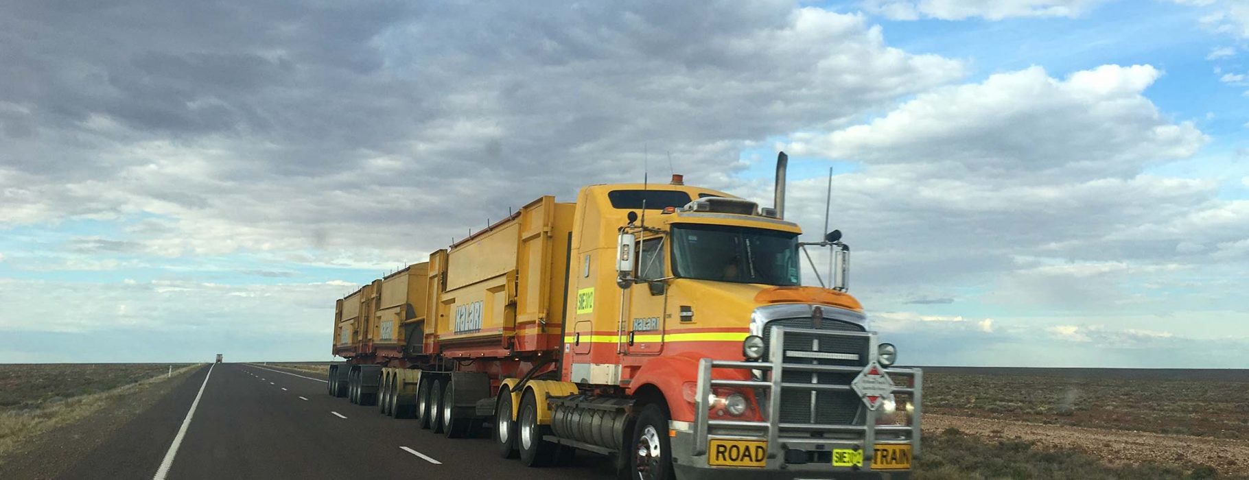 A lone truck or "road train" travels down an otherwise empty Australian road, beneath a blue and party cloudy sky